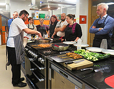 Cookery class with people standing around watching chef cook in a pan on stove