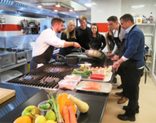 group of men and women surrounding a cooking unit whilst watching a chef lean forward swirling oil in a pan