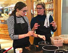 Lady talking to another lady whilst the girl listening is stirring a spoon in a pan on a stove